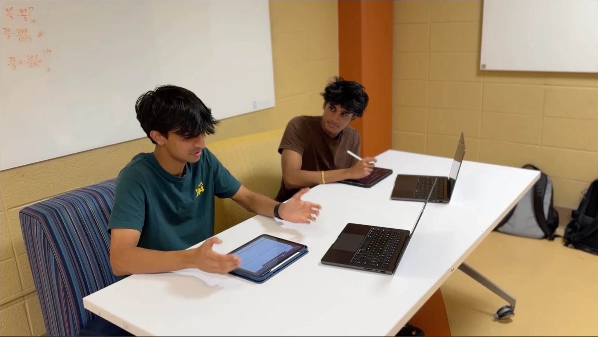Two students working through a problem together at a table with laptops and iPads.