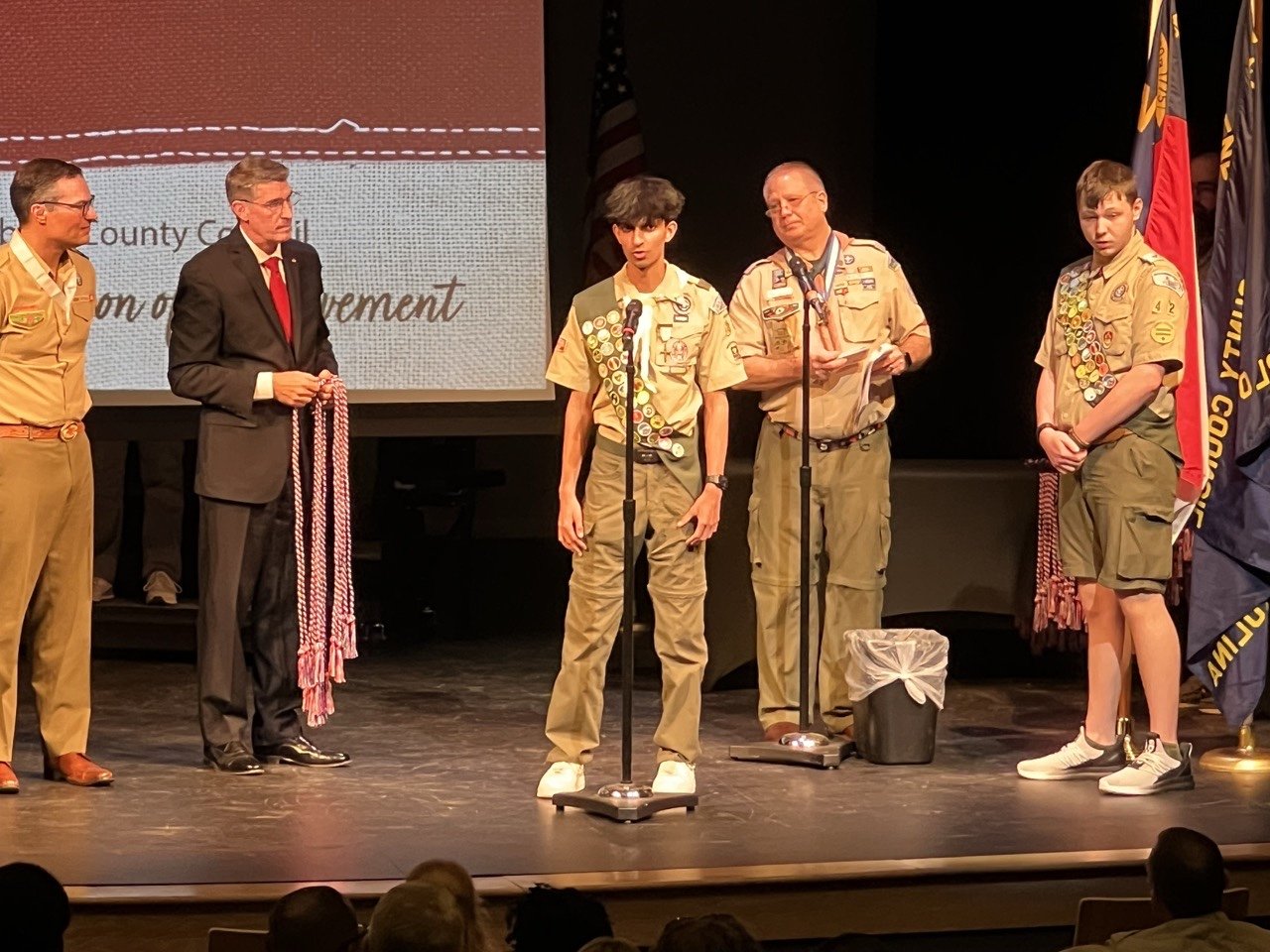 Jason in Scout uniform speaking at a microphone on stage, with Scout leaders and officials beside him.